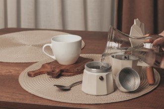 Hot water is being poured from a glass kettle into a coffee maker on a wooden table. A white cup