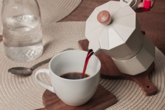 Freshly brewed coffee is being poured from a stovetop espresso maker into a white cup on a wooden