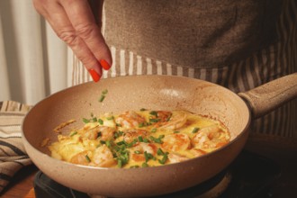 A woman is cooking shrimp in a skillet, adding fresh herbs to the dish. The warm colors of the