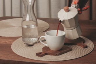 A hand pours freshly brewed coffee from a stovetop maker into a white cup. A glass pitcher filled
