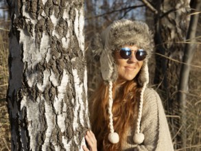 Slender pretty woman with fur hat and pompoms, red hair and warm winter colors between birch forest