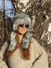 Slender pretty woman with Mongol hat, red hair and warm winter colors between birch forest and