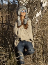 Slim woman with fur hat and sunglasses, red hair and warm winter colors between birch forest and