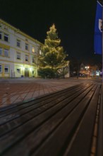 A brightly lit Christmas tree next to a building and a wooden bench, Aidlingen, Böblingen district,