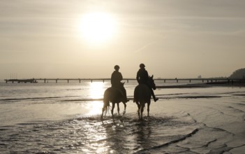 Two female riders ride their horses through the shallow water of the Baltic Sea at sunrise,
