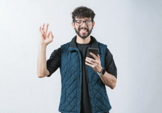 Smiling handsome man holding phone and gesturing OK, isolated. Attractive man in glasses holding