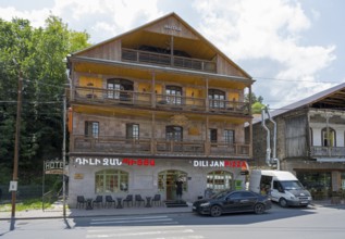 Three-story hotel with wooden faÃ§ade and pizzeria, surrounded by trees and cars on the street,