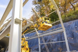 Workers in a yellow lift work on a raised metal structure in front of a forest, Hermann Hesse