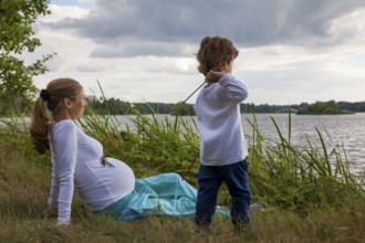 Pregnant woman with little boy at the lake, in grass, child playing with sticks