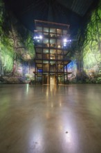 A wooden tower stands in front of a green illuminated artificial jungle backdrop, Amazonia