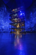 An illuminated, multi-storey structure in front of a blue artificial forest projection, Amazonia