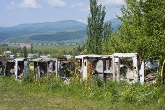 Row of rusty cars in a meadow with green trees and mountains, scrap cars serving as fencing near
