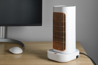 A sleek electric heater stands on a wooden desk next to a computer monitor. The heater features a