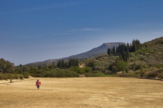 A child runs across a dry meadow to distant mountains, under a clear sky with cypresses in the