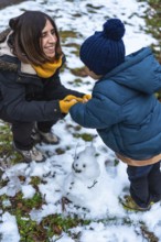Happy mother and young son build a small snowman outdoors, laughing and bonding in warm parkas,