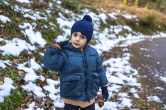 Young boy in warm parka and hat holding a snowball, smiling while playing in a snowy forest