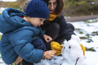 Mother and young son crouch in fresh snow, building and adding details to a tiny snowman while
