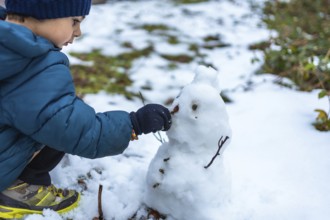Young boy playing in the snow, carefully placing a small natural element on a snowman he is