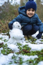 Smiling young boy crouching outdoors, happily creating a small snowman on a patch of grass lightly