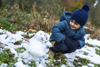 Young boy kneeling on snowy ground, wearing a warm coat, hat, and gloves, observing a freshly made