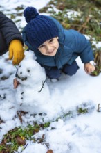 Young child wearing a blue knitted hat and warm winter clothes kneeling in the snow, proudly