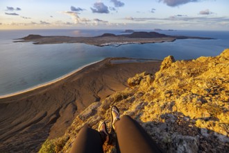 First person view, legs of a woman sitting on a steep cliff, view of the sea and the island of La