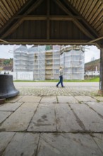 A woman walks under an arched roof, in the background a building with scaffolding in a village,