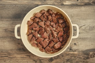 Chunks of beef are searing in a large pot on a weathered wooden table. The meat is cooked to a rich