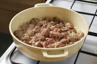 Pieces of beef are sizzling and cooking in a large pot on a gas stove. The meat is browning while