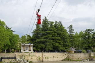 A red cable car cabin above tree tops near a river under a cloudy sky, cable car station, Kutaisi,