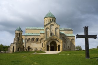 Historic church under a cloudy sky surrounded by greenery with a large cross in the foreground,