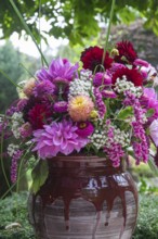 A vivid bouquet of flowers in shades of pink and red stands in a rustic vase against a green