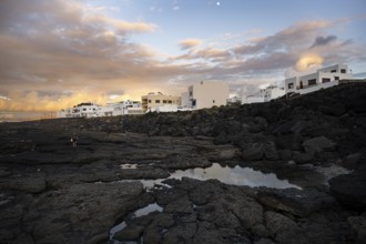Black coast of volcanic rocks behind typical white houses of the village of La Santa, at sunset,