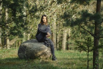 A young woman is seated on a large rock in the midst of a vibrant green forest filled with blooming