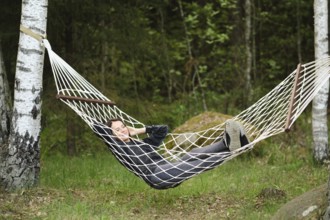 A woman enjoys a peaceful afternoon in a hammock, nestled between trees. She has a relaxed posture,