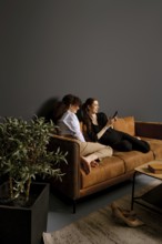 Two friends sit comfortably on a brown couch in a modern living room, engaged with a mobile device.