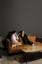 Two friends sit on a comfortable couch in a stylish living room. One is scrolling on a smartphone