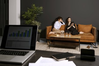 Two friends sit comfortably on a brown couch in a stylish living room, engaging with a smartphone.