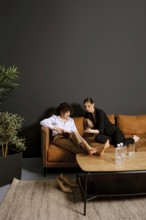 Two friends sit comfortably on a warm brown couch in a stylish room, engaging in conversation. Lush