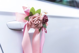 Elegant pink ribbons and artificial flowers adorn the handle of a white car, signifying a wedding