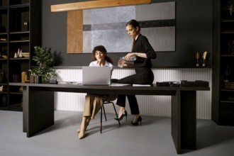 In a stylish office, two women engage in a collaborative discussion. One woman sits at a sleek