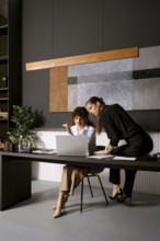 Two women are working together in a stylish office. One woman sits at a table with a laptop while