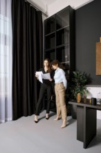 Two professional women stand in a sleek office, reviewing important documents. Sunlight filters