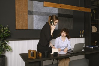 Two women are engaged in a work discussion in a stylish office setting. One is seated at a desk