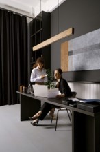 Two businesswomen are engaged in a discussion at a sleek office desk. One is seated while reviewing