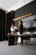 Two women are engaged in a discussion in a contemporary office space, surrounded by dark walls and