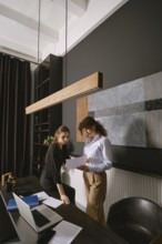 Two women engaged in conversation while reviewing documents in a stylish office. The setting