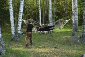 Two children spend a sunny afternoon in a forest, one relaxing in a hammock strung between birch