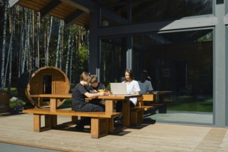 Two children focus on their tasks at a wooden table outside a modern cabin, while their mother