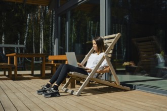 A woman sits comfortably in a wooden chair on a deck, focused on her laptop. Bright sunlight